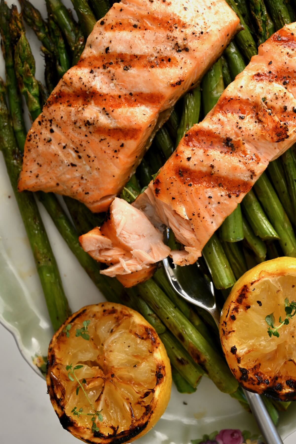 Two grilled salmon fillets on a bed of grilled asparagus, with one fillet flaked with a fork and two grilled lemon slices on the side. - 1