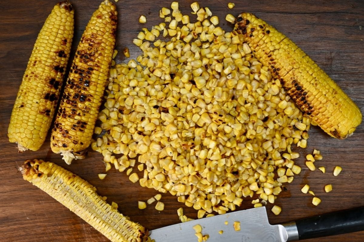 A grilled corn cob being cut into kernels on a wooden board.