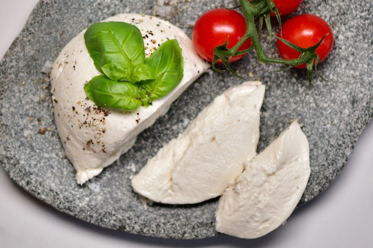 Vegan mozzarella cheese block with two slices cut, served on a plate with tomatoes on the side and basil leaves on top.