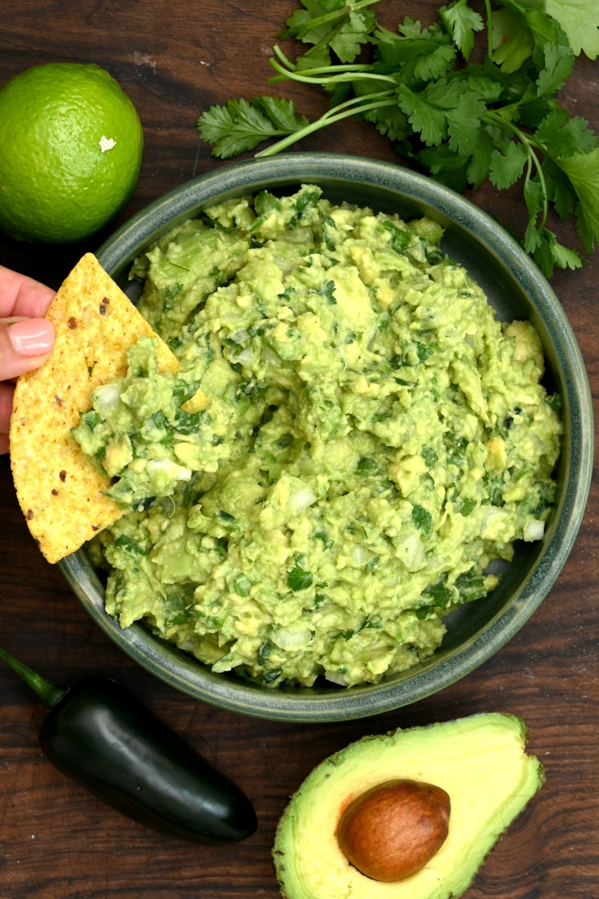 Vertical top view of guacamole in a green bowl with a tortilla chip held above it, surrounded by a halved avocado, jalapeño, lime, and cilantro on a wooden board.