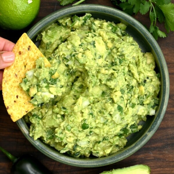 Top view of guacamole in a green bowl with a tortilla chip dipped into it, surrounded by lime, cilantro, avocado, and jalapeño on a wooden board.
