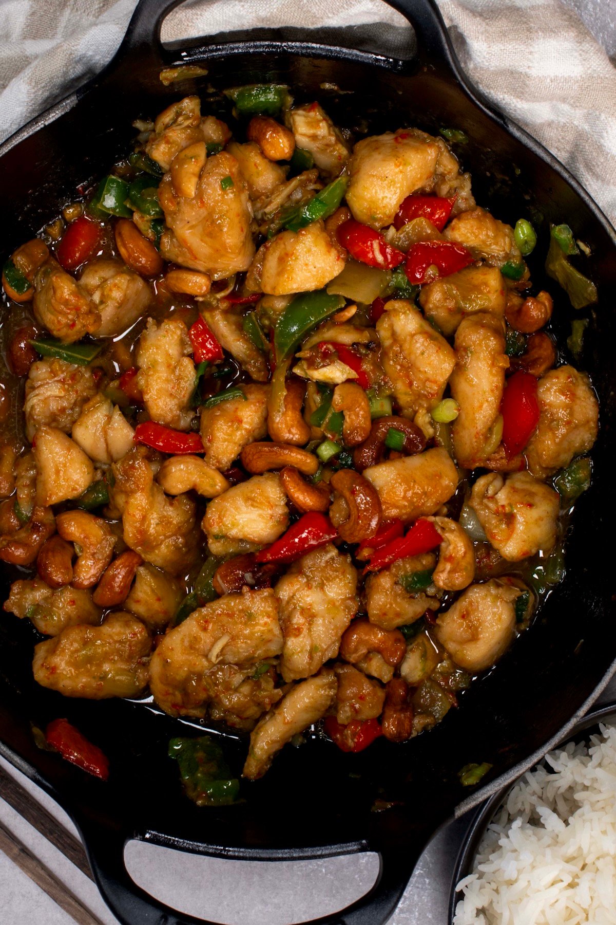 Top view of cashew chicken stir-fry in a black wok with a bowl of white rice beside it.