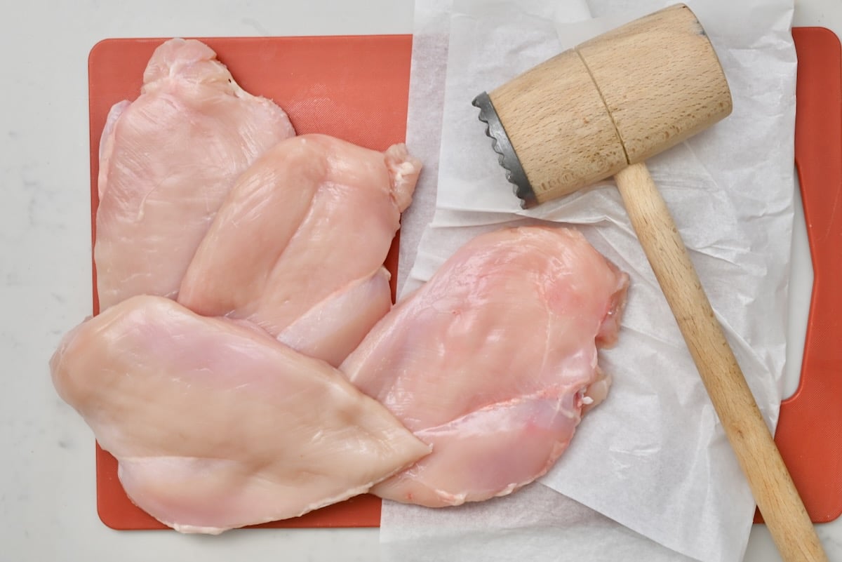 1 Raw pounded chicken breasts on an orange cutting board with a wooden meat mallet beside them, top view