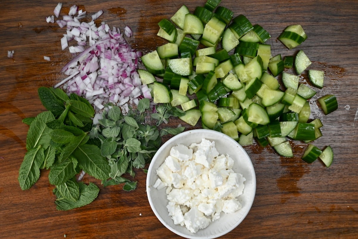 Top view of chopped cucumbers, red onions, fresh mint and oregano leaves with a small bowl of crumbled feta, all arranged on a wooden board.