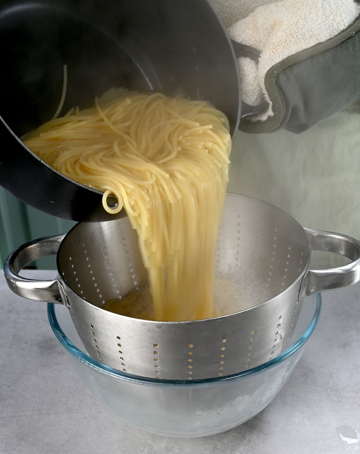 Side view of me draining cooked spaghetti in a colander with a glass underneath to catch pasta water.