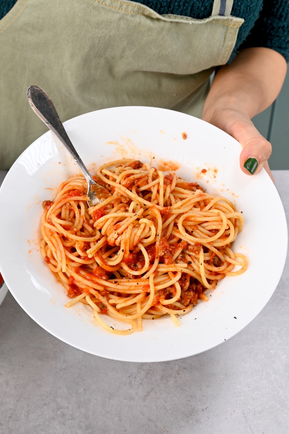 Side view of a white plate of spaghetti with sauce, held in one hand with a fork twirling pasta.