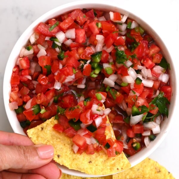 Close-up top view of pico de gallo in a white bowl, with tortilla chips and pico de gallo held by hand.