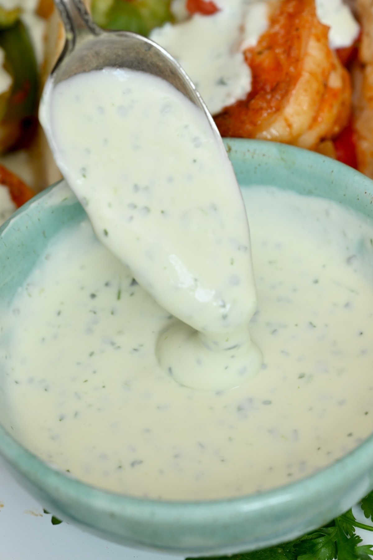 Side view close-up of lime crema in a green bowl with a spoon, shrimp fajitas faintly visible in the background.