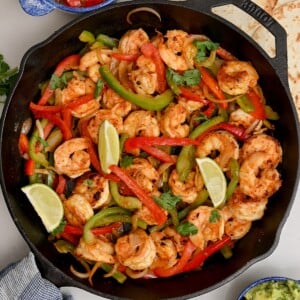 Top view of shrimp fajitas in a black cast iron skillet with lime wedges, on a square frame.