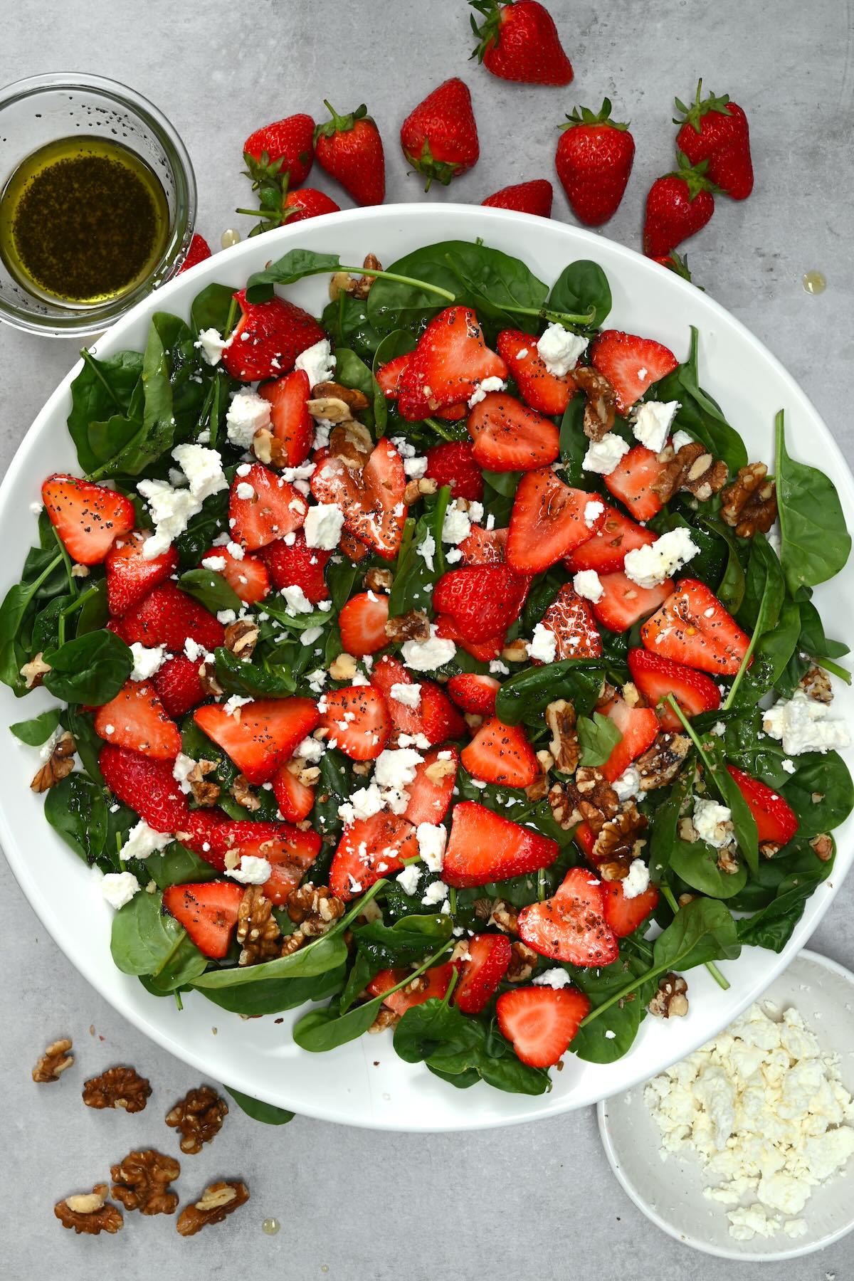 Top view of strawberry spinach salad on a grey background, with strawberries, dressing, walnuts, and crumbled feta cheese scattered nearby.
