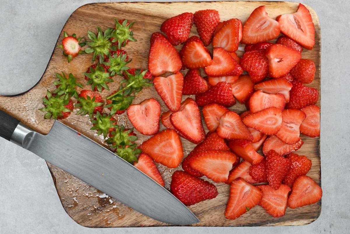 Top-down view of sliced strawberries on a wooden board, with strawberry tops and a knife beside them.