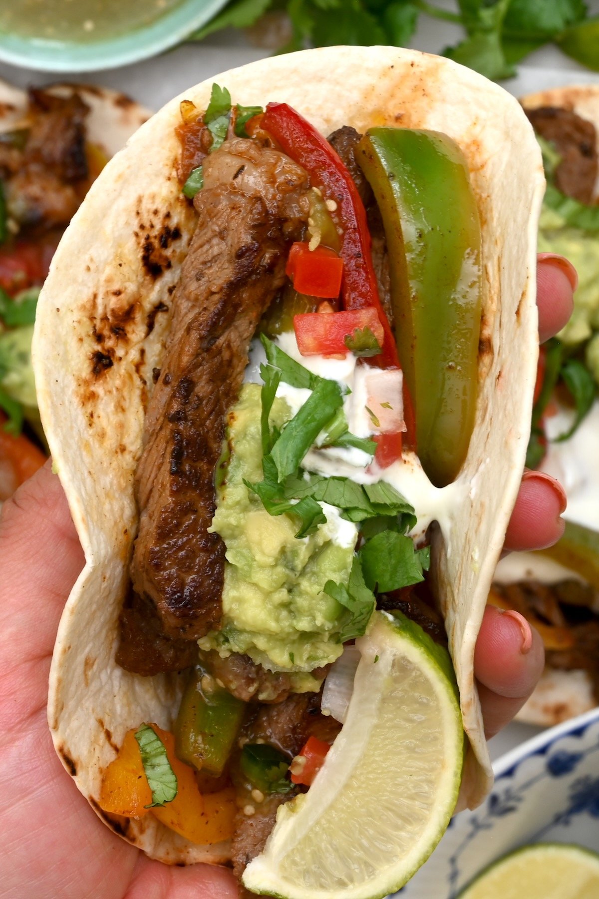 Top view close-up of a steak fajita in a small tortilla with lime wedge, sour cream, guacamole, and cilantro.