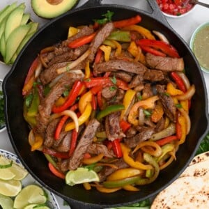 Top view of steak fajitas in a black cast iron skillet, square photo with garnishes on the side.