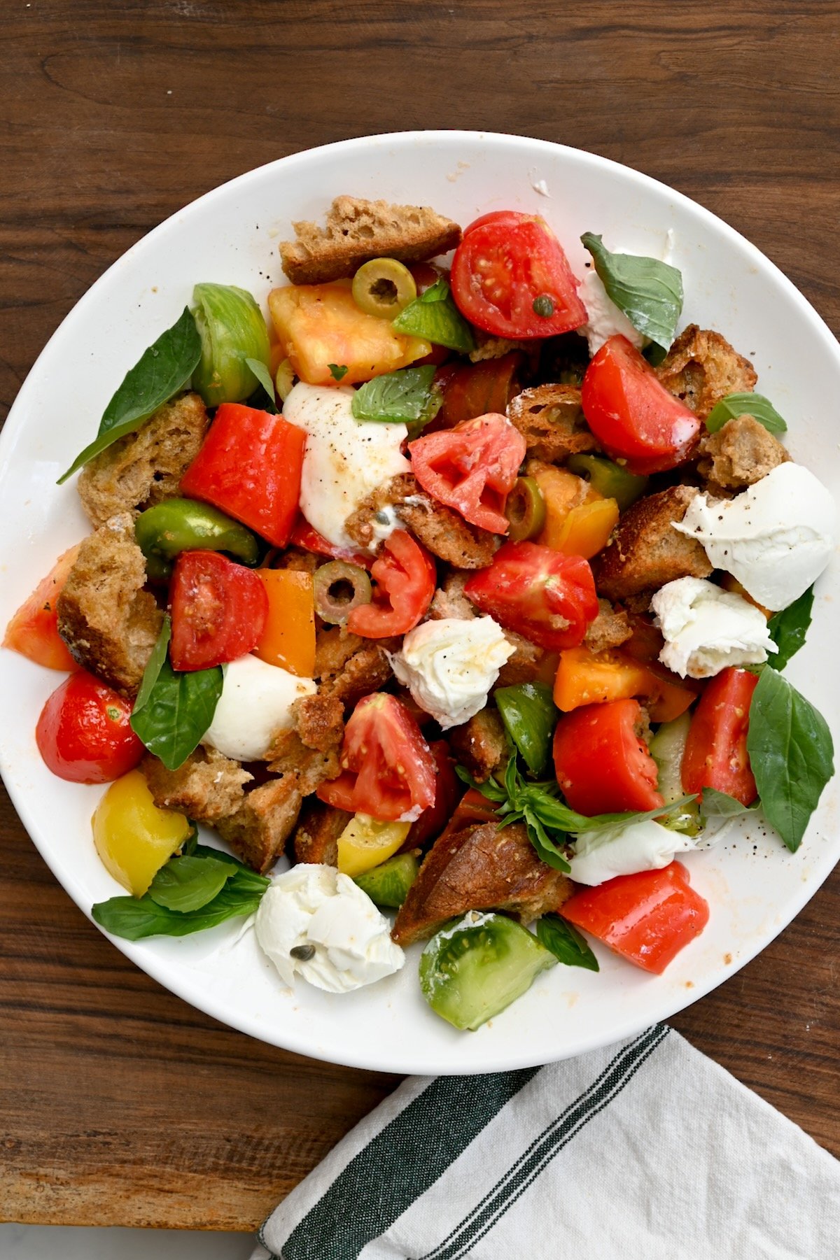 Vertical top view of panzanella salad in a white bowl on a wooden background with an off-white cloth beside it.