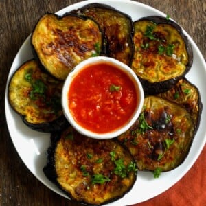 Top view of roasted eggplant slices arranged on a white dish with a bowl of sauce in the center.