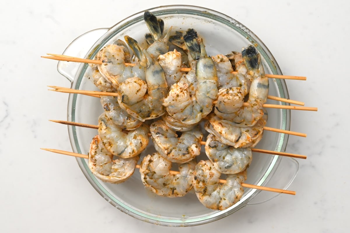 Shrimp skewers over glass bowl – Top view of six raw shrimp skewers resting across a glass bowl on white marble.