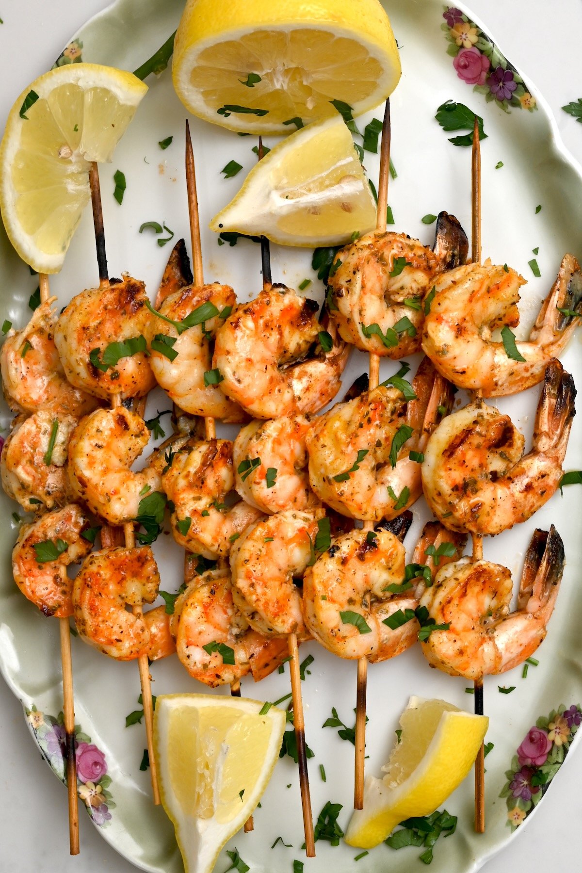 Shrimp skewers with garnish – Top view of six grilled shrimp skewers on a green plate garnished with parsley and lemon wedges.