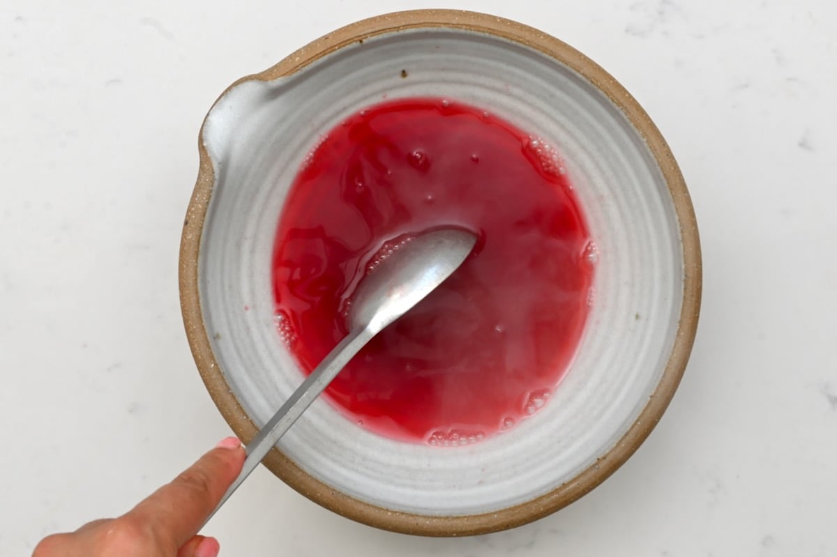Stirring strawberry Jello with warm water in a gray bowl with a brown rim, on white marble. - 6