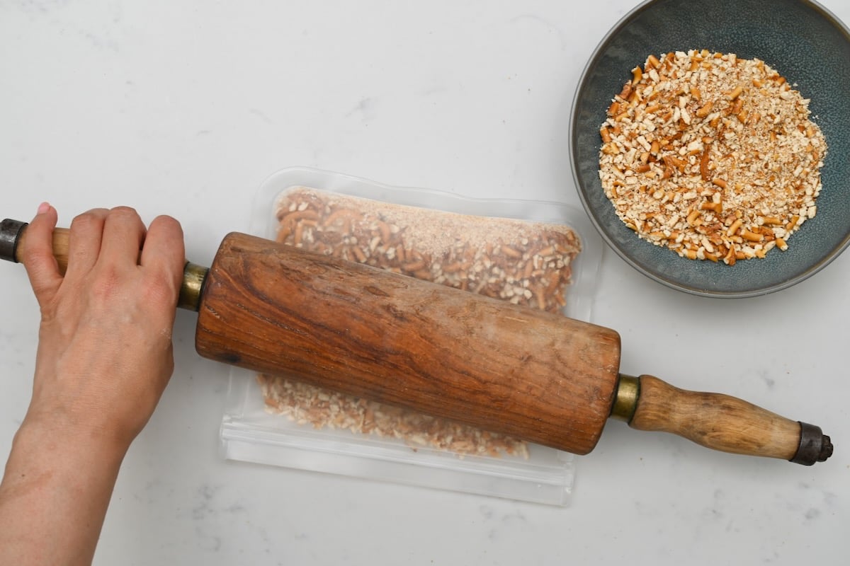 Top-down view of pretzels in a Ziploc bag with a rolling pin above, next to a blue plate of crushed pretzels on white marble. - 3