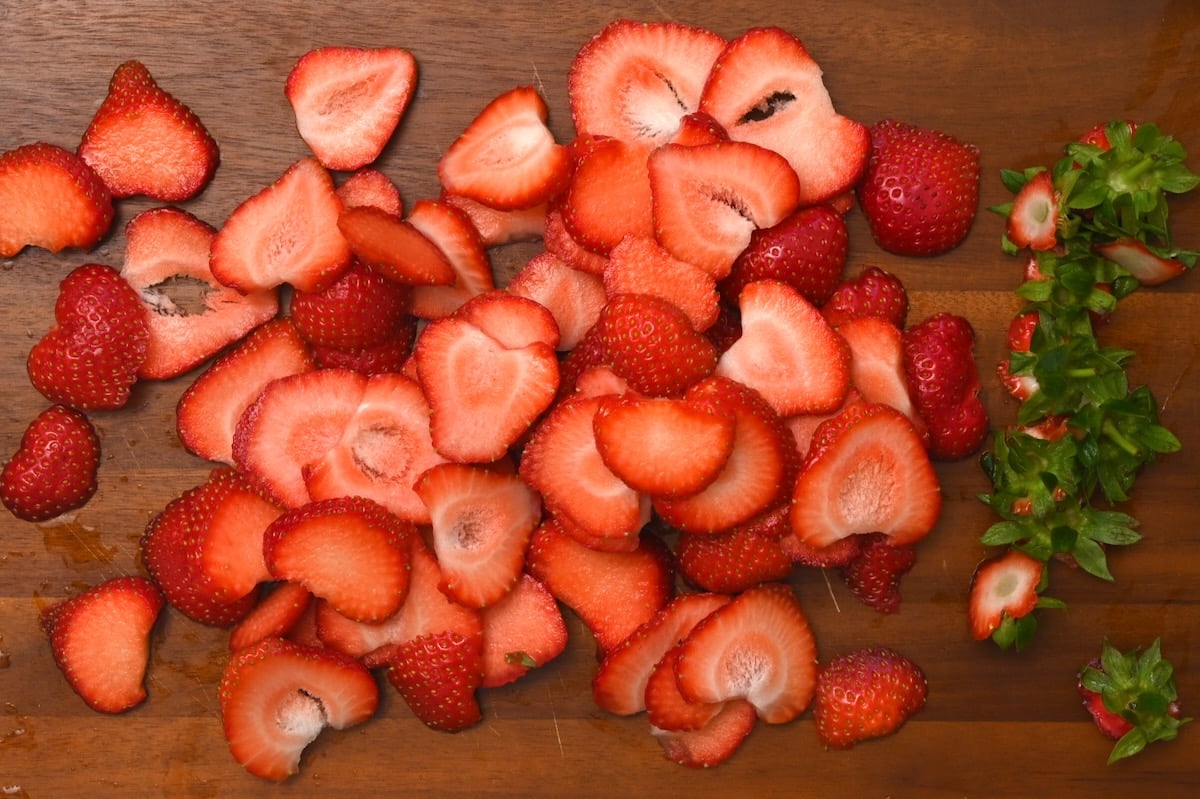 Sliced strawberries on a wooden board with strawberry tops set aside. - 5