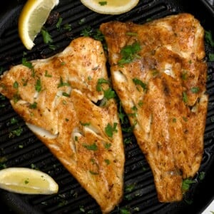 top view of grilled fish in a black grill pan with parsley sprinkle and lemon wedges.
