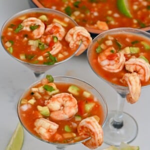 side view of three mexican shrimp cocktail glasses with large bowl in the background and lime wedges