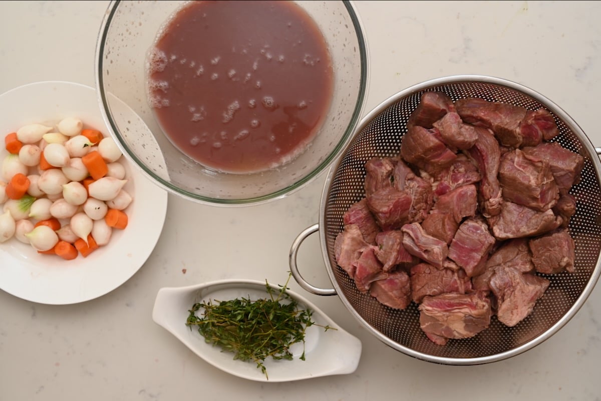 Strained red wine marinade, drained beef pieces, pearl onions, carrots, and thyme laid out after marinating for beef bourguignon. - 9