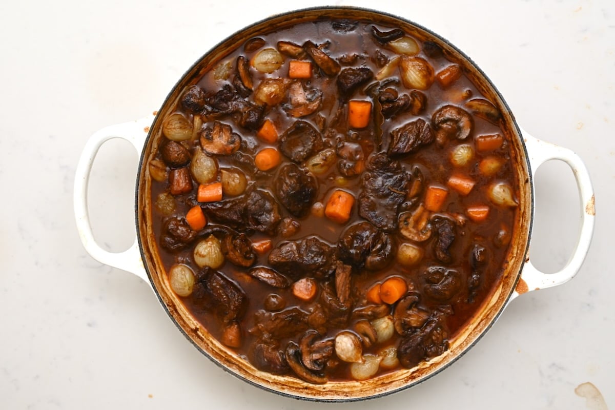 A pot of finished beef bourguignon simmering with tender beef, carrots, mushrooms, and pearl onions in a rich red wine sauce. - 24
