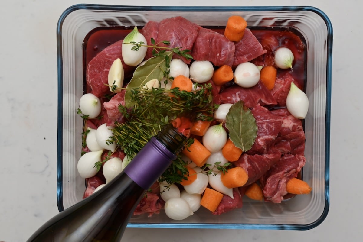 Red wine being poured over beef bourguignon ingredients including beef chunks, carrots, pearl onions, thyme, and bay leaves. - 6