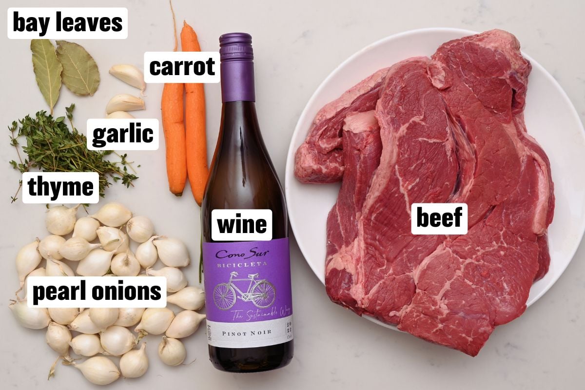 Beef, red wine, pearl onions, carrots, garlic, thyme, and bay leaves laid out on a counter. - 2