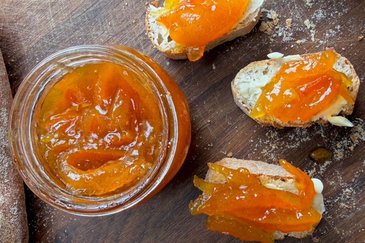 Jar of homemade orange marmalade served with sliced bread spread with butter and marmalade on a wooden board.” - 7