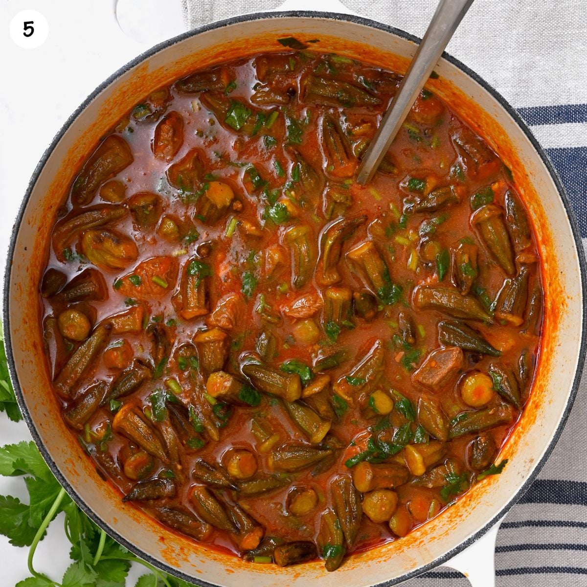 Close-up of the finished bamya (okra) stew in a pot, showing tender okra in a rich tomato sauce, with fresh green cilantro being sprinkled on top.
