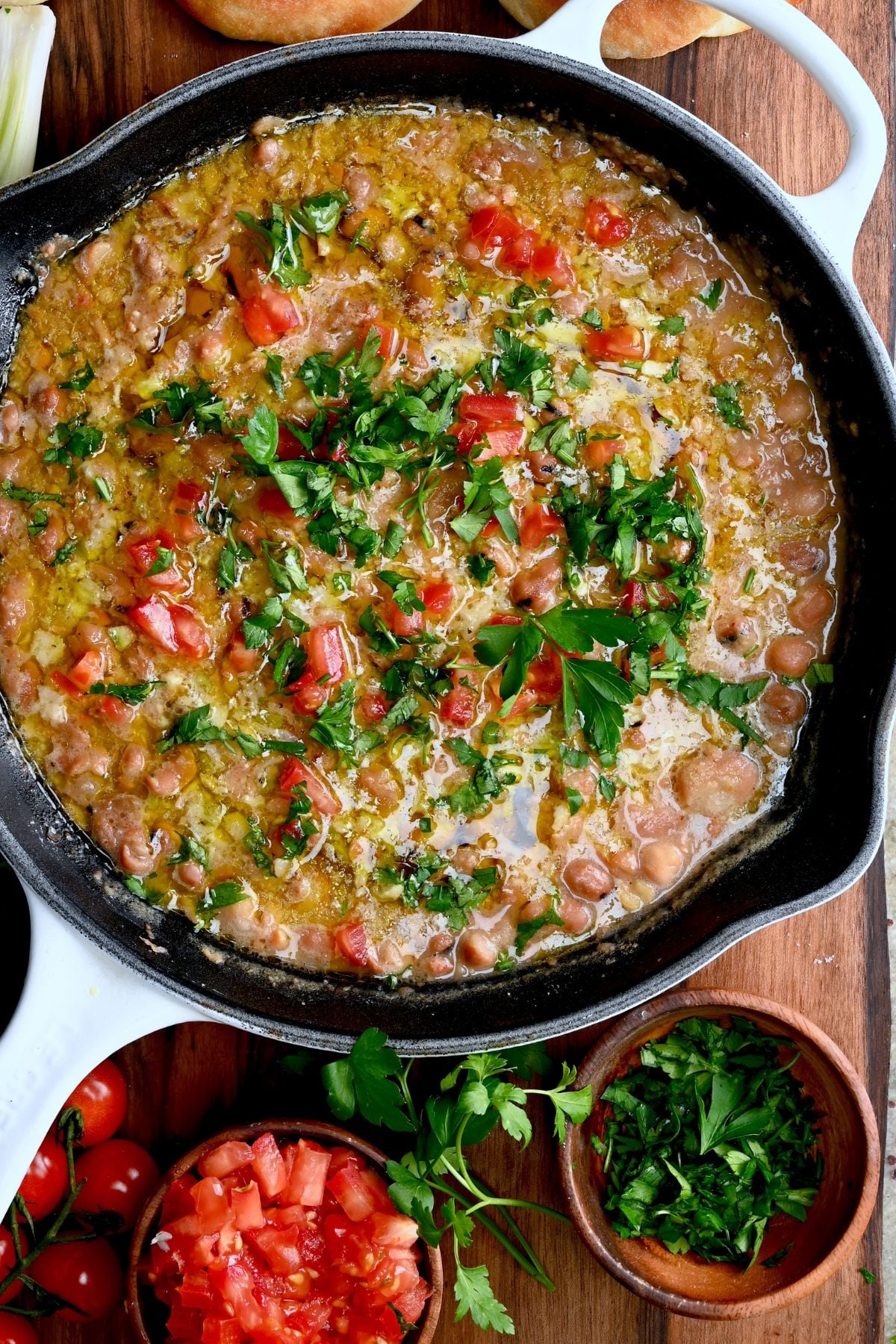 Ful medames in a skillet, topped with fresh parsley and tomatoes and drizzled with olive oil.