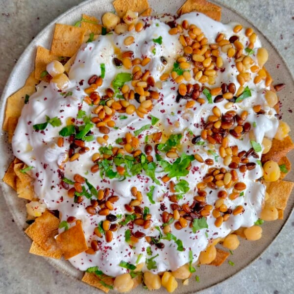 Overhead view of chickpea fatteh with crispy pita, creamy yogurt sauce, toasted pine nuts, and chopped parsley.