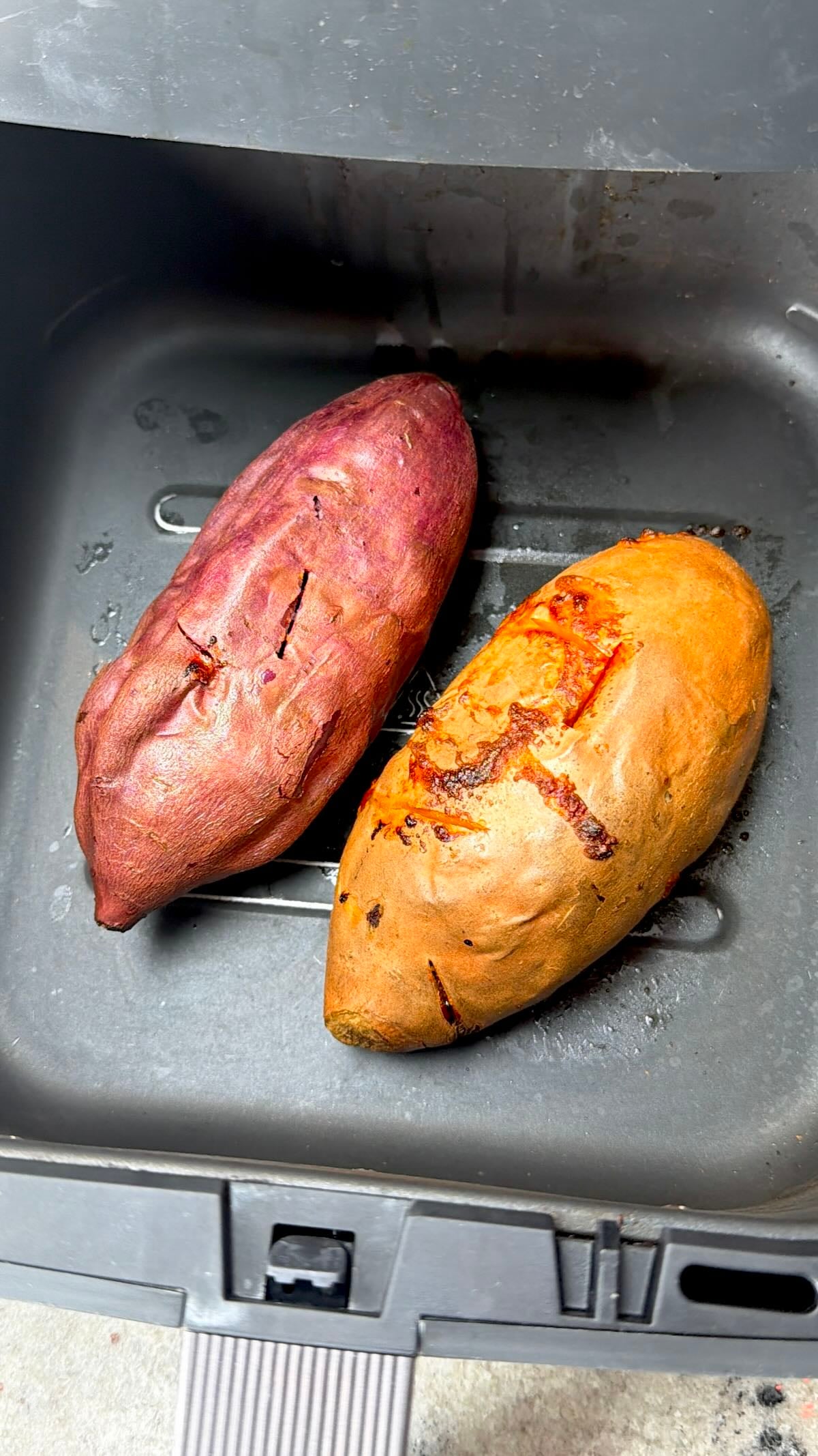 Two whole sweet potatoes (purple and orange) cooking in an air fryer basket.