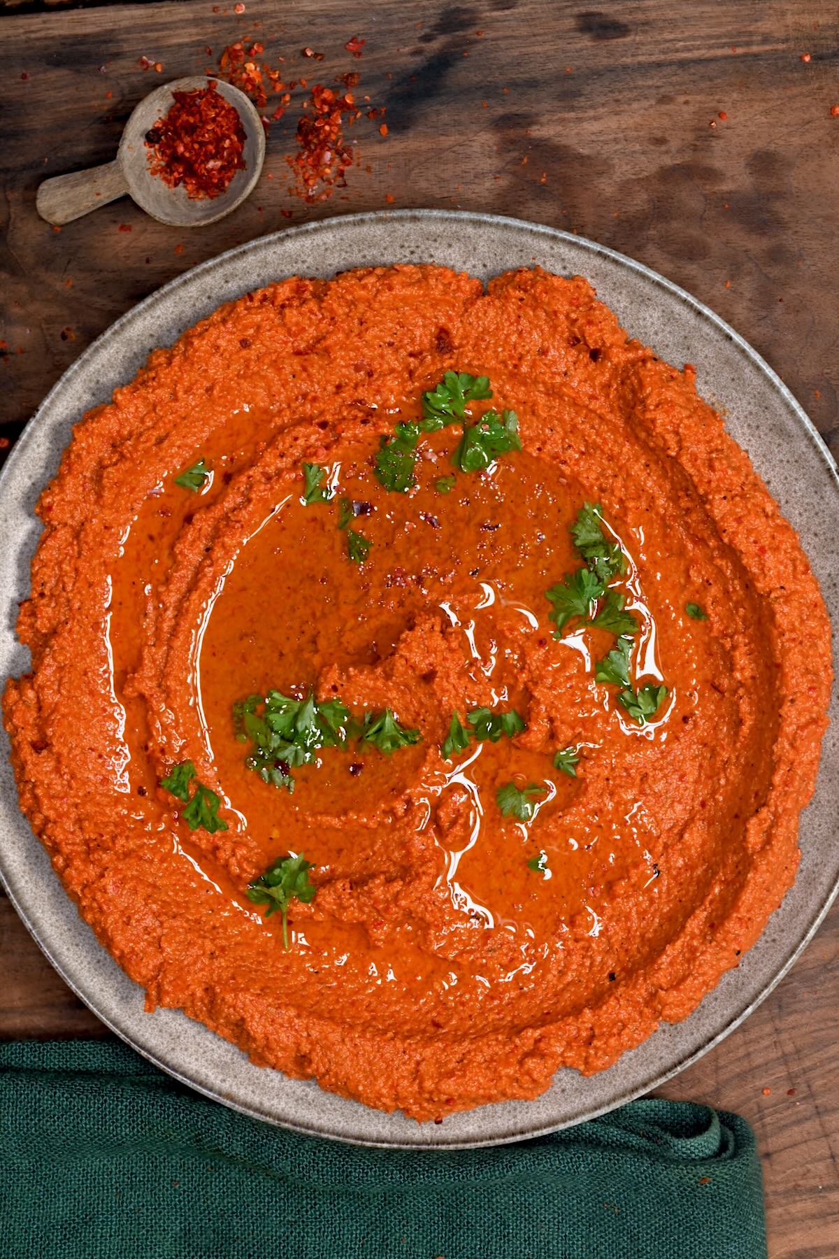 A top-down view of a creamy bowl of authentic Muhammara, a red pepper and walnut dip, swirled with olive oil and garnished with fresh parsley. A small wooden spoon filled with red pepper flakes sits on the rustic wooden table alongside.