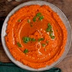 A close-up of a rustic plate of vibrant, orange-red Muhammara dip topped with a generous swirl of olive oil and chopped green parsley. The texture shows the finely ground toasted walnuts and roasted red peppers.