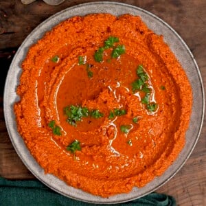 A close-up of a rustic plate of vibrant, orange-red Muhammara dip topped with a generous swirl of olive oil and chopped green parsley. The texture shows the finely ground toasted walnuts and roasted red peppers.