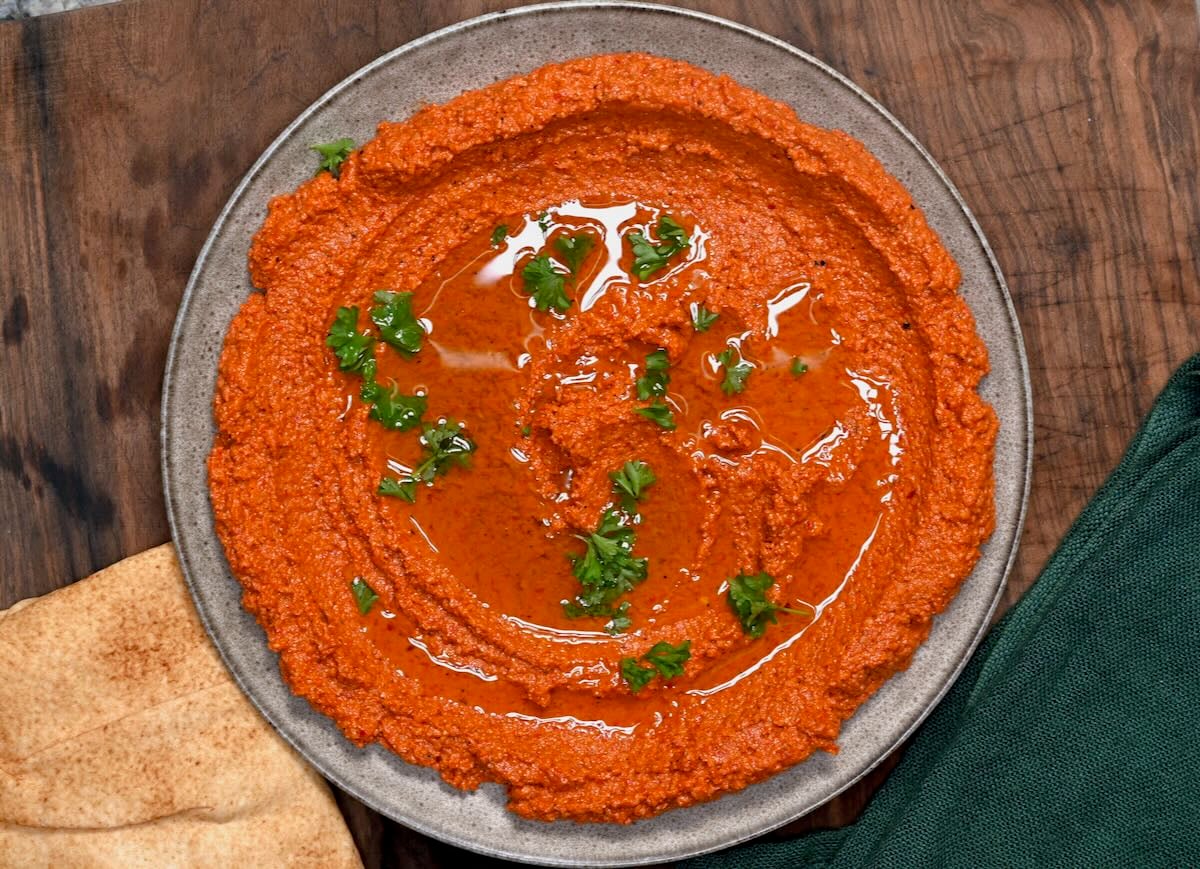 Muhammara dip served on a plate and topped with olive oil and parsley with pita next to it.