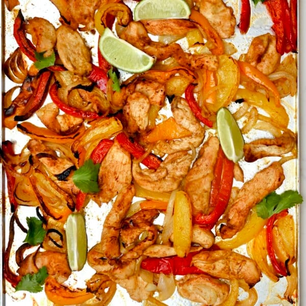 square photo of sheet pan of baked chicken strips, red and yellow peppers, and onions with lime wedges and cilantro.