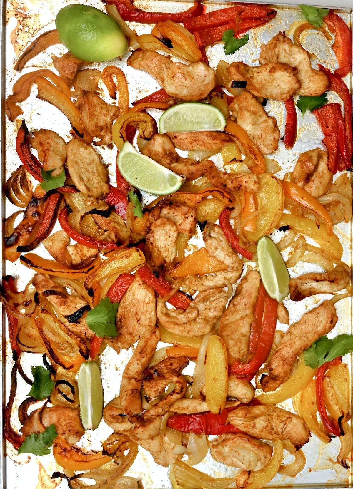 Sheet pan of baked chicken strips, red and yellow peppers, and onions with lime wedges and cilantro.