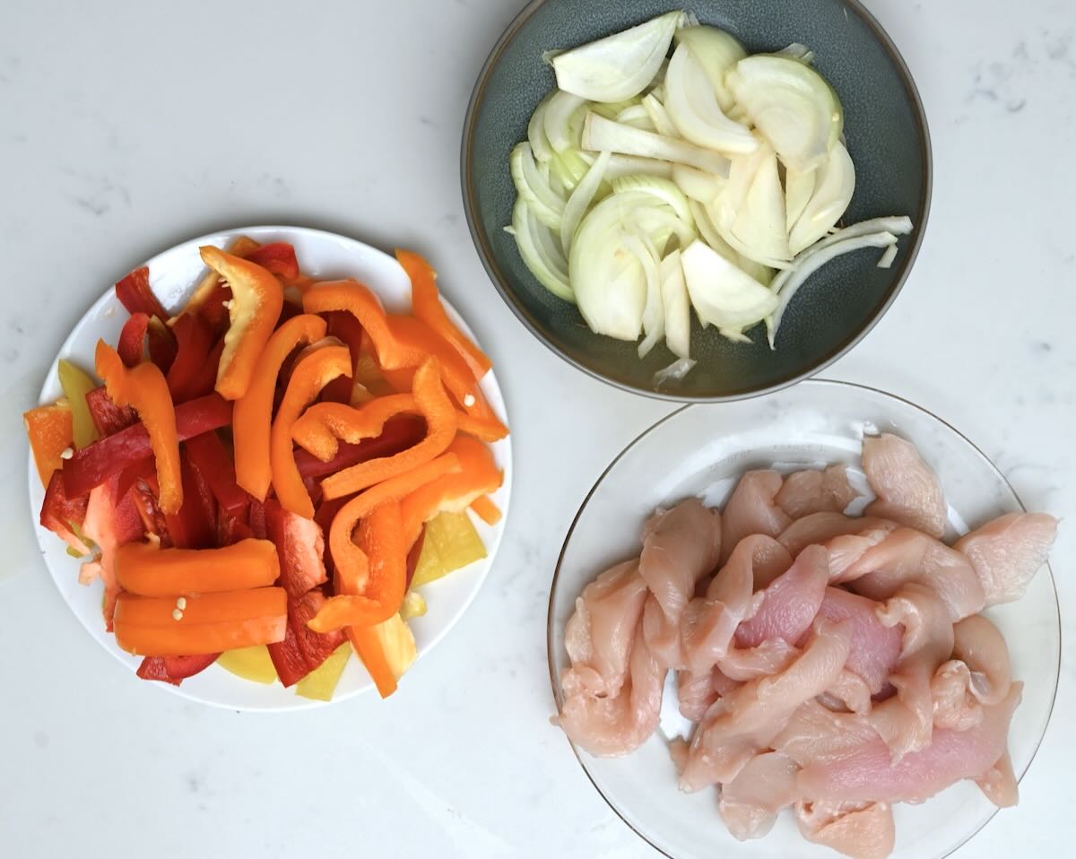 Three plates showing prepped ingredients for sheet pan chicken fajitas, sliced raw chicken breast, sliced bell peppers, and sliced onion