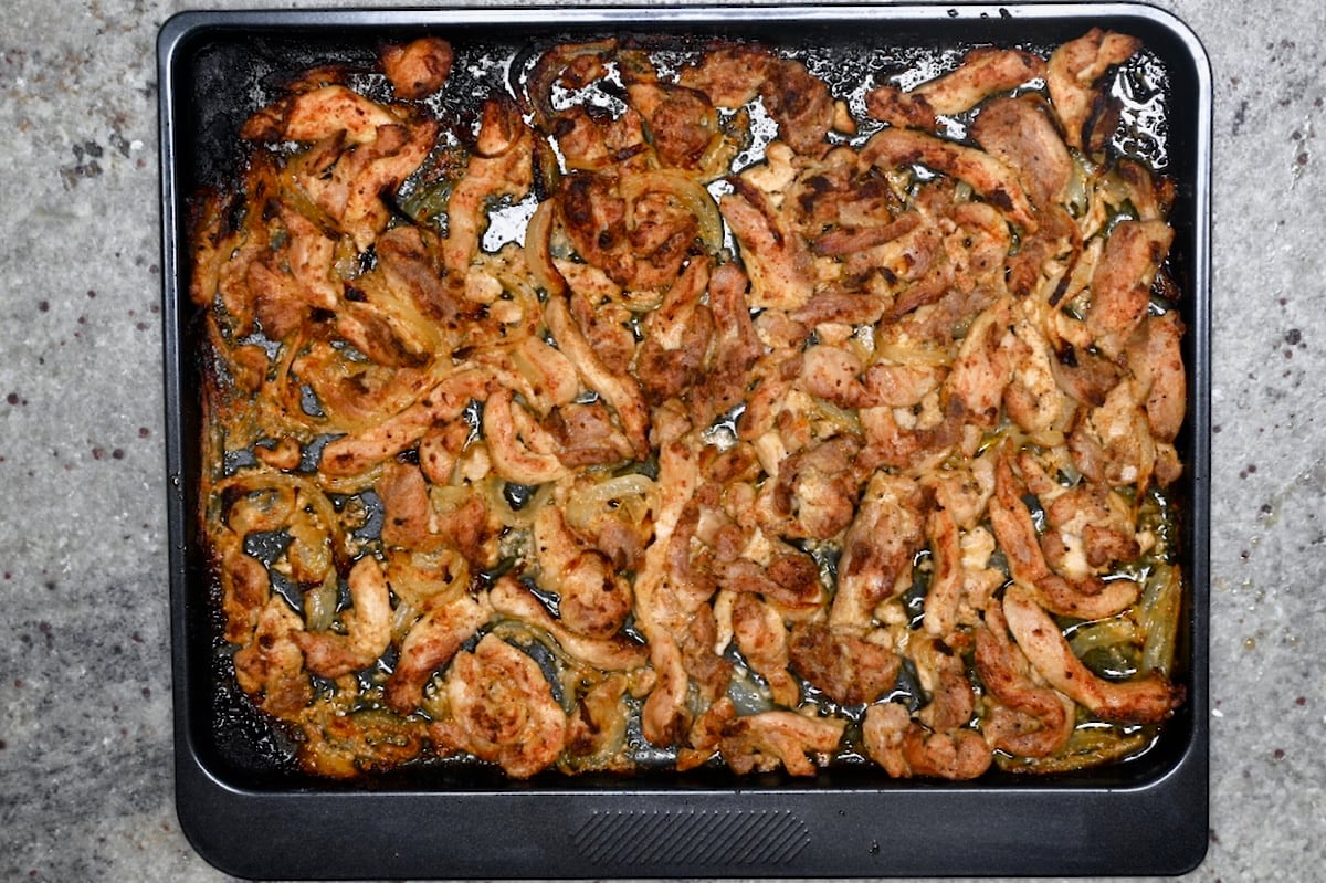 Baked chicken shawarma strips and onions with browned edges on a sheet pan.