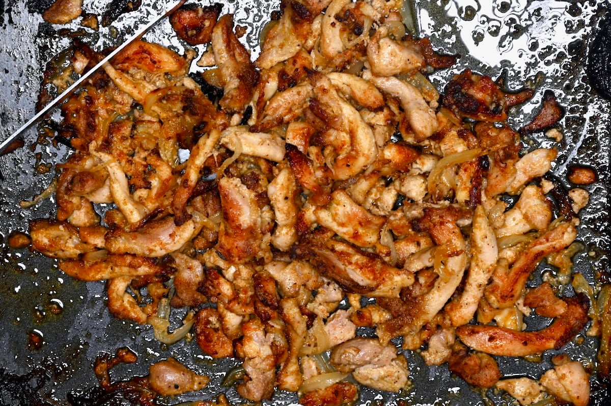 Close-up of cooked chicken shawarma strips and onions with charred bits on a pan.