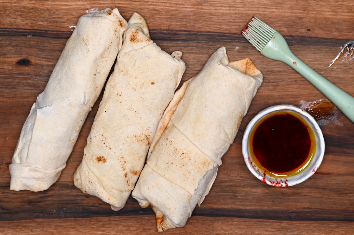Three wrapped chicken shawarma wraps on a wooden board with a pastry brush and spiced oil.