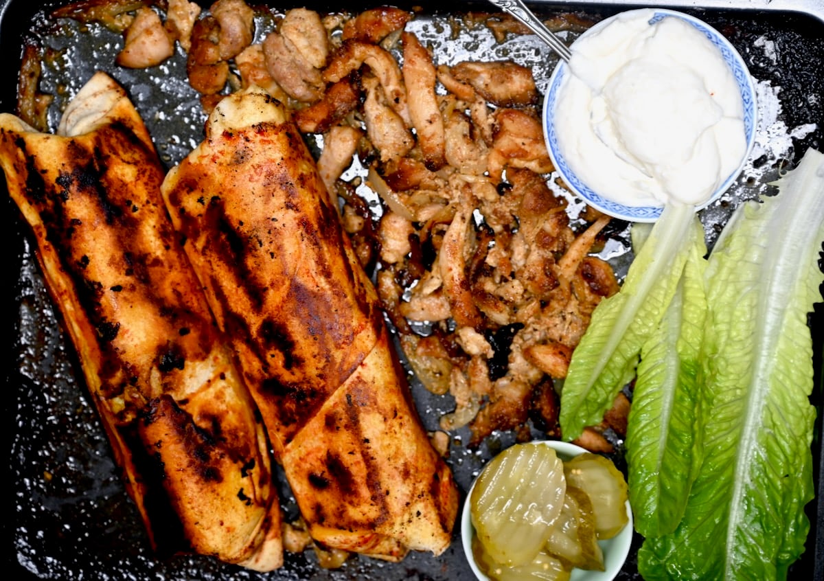 Serving tray with crispy shawarma wraps, cooked chicken, garlic sauce, pickles, and romaine lettuce.