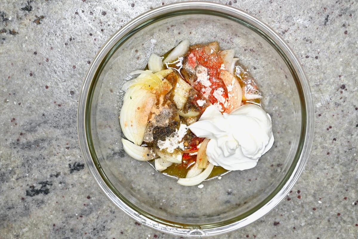 Overhead view of shawarma marinade ingredients in a glass bowl with yogurt, sliced onion, lemon wedges, and spices.