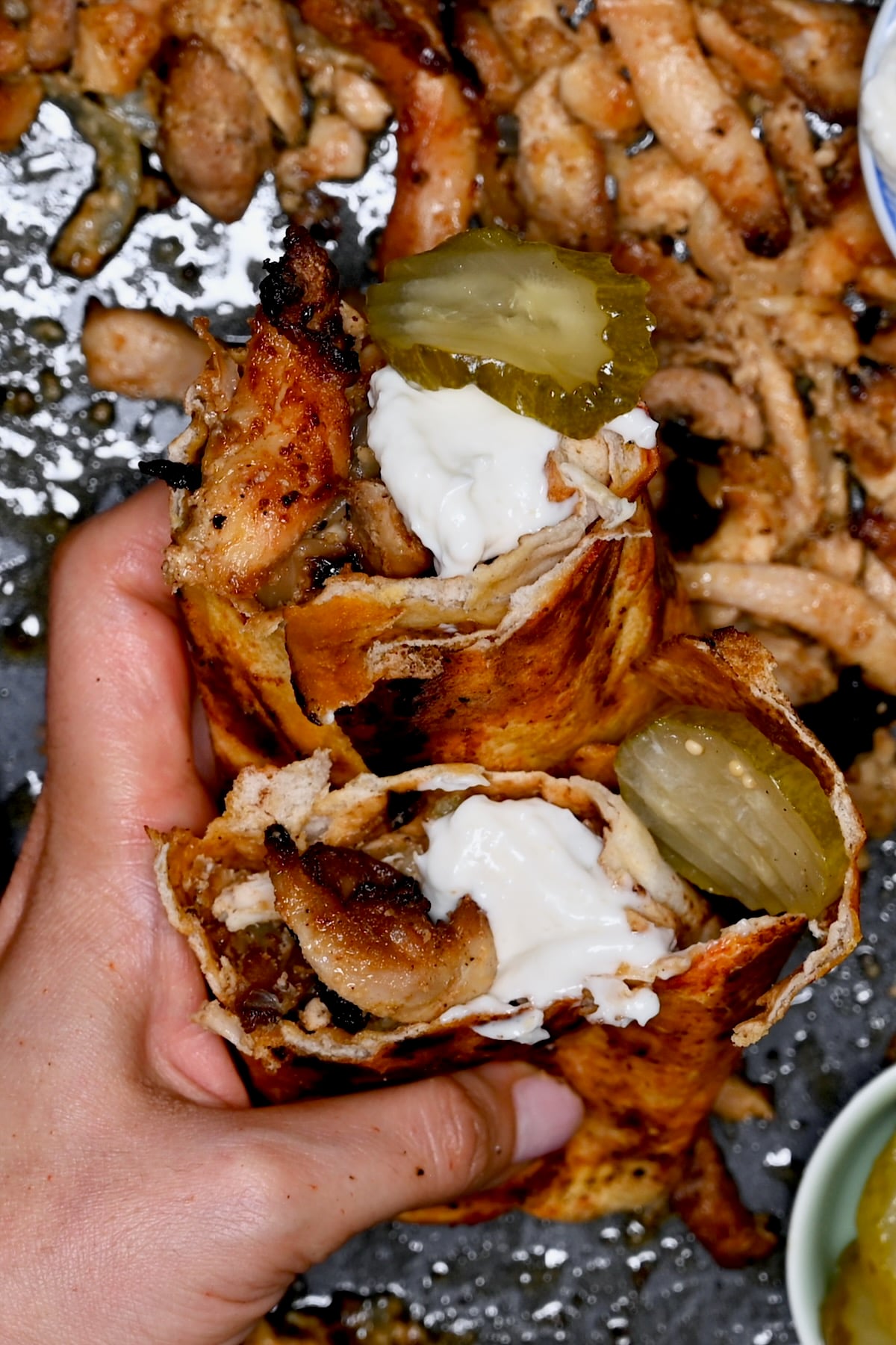 Hand holding halved chicken shawarma wrap with garlic sauce and pickle slices.