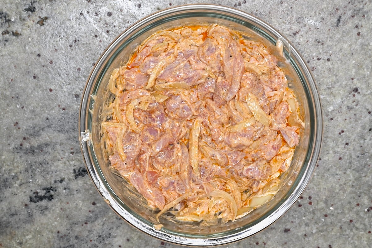 Chicken strips and onions fully coated in shawarma yogurt marinade in a glass bowl.