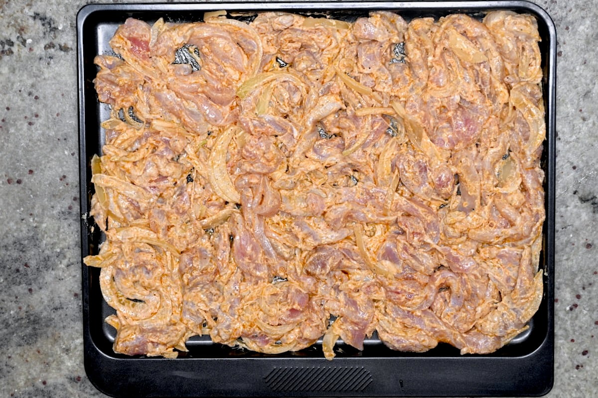 Marinated chicken shawarma and onions spread on a baking sheet before cooking.